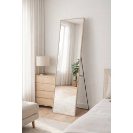 Lustro stojące w sypialni o czarnych ramkach, nachylone przy ścianie, obok drewnianej komody z lampą i zasłonami.